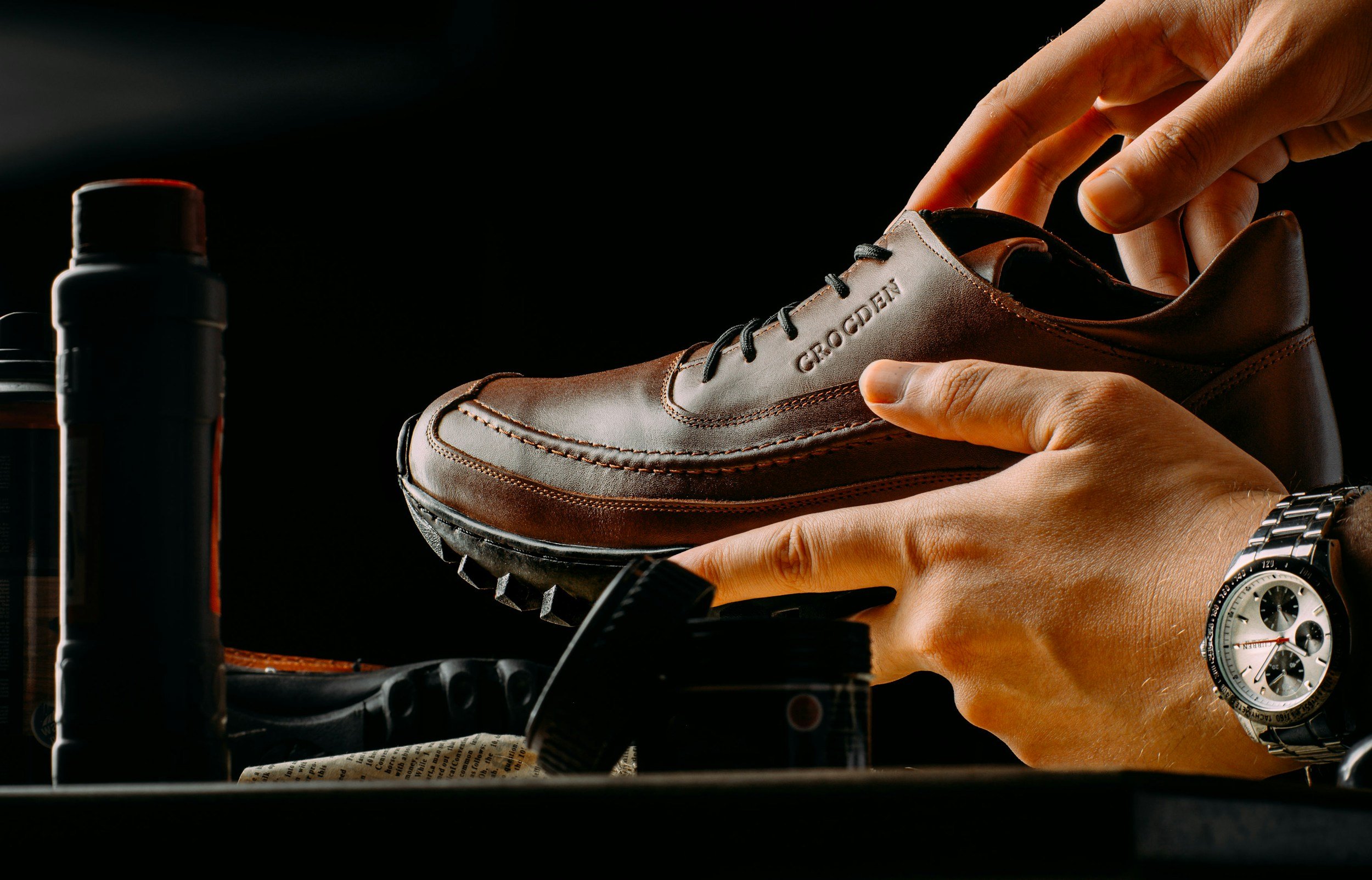 Person polishing a brown leather shoe labeled 'GRODEN' with sneaker shoe polish, wearing a silver wristwatch.