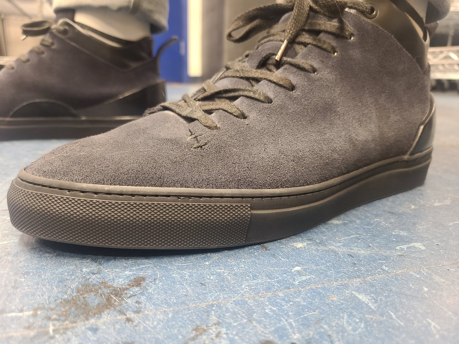 A gray suede sneaker with black rubber sole on a blue floor, with another dark sneaker in the background.