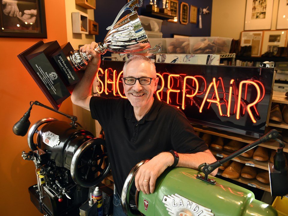 Smiling man wearing glasses inside a shoe repair shop, holding a large trophy above his head, with a neon sign that reads 'SHOE REPAIR' and shoe repair equipment around him.
