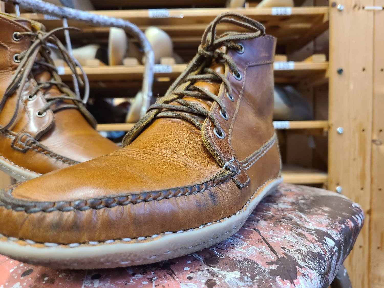 Close-up of a worn, tan leather moccasin-style shoe with visible stitching, placed on a weathered surface, with wooden shelves and other shoes in the background.
