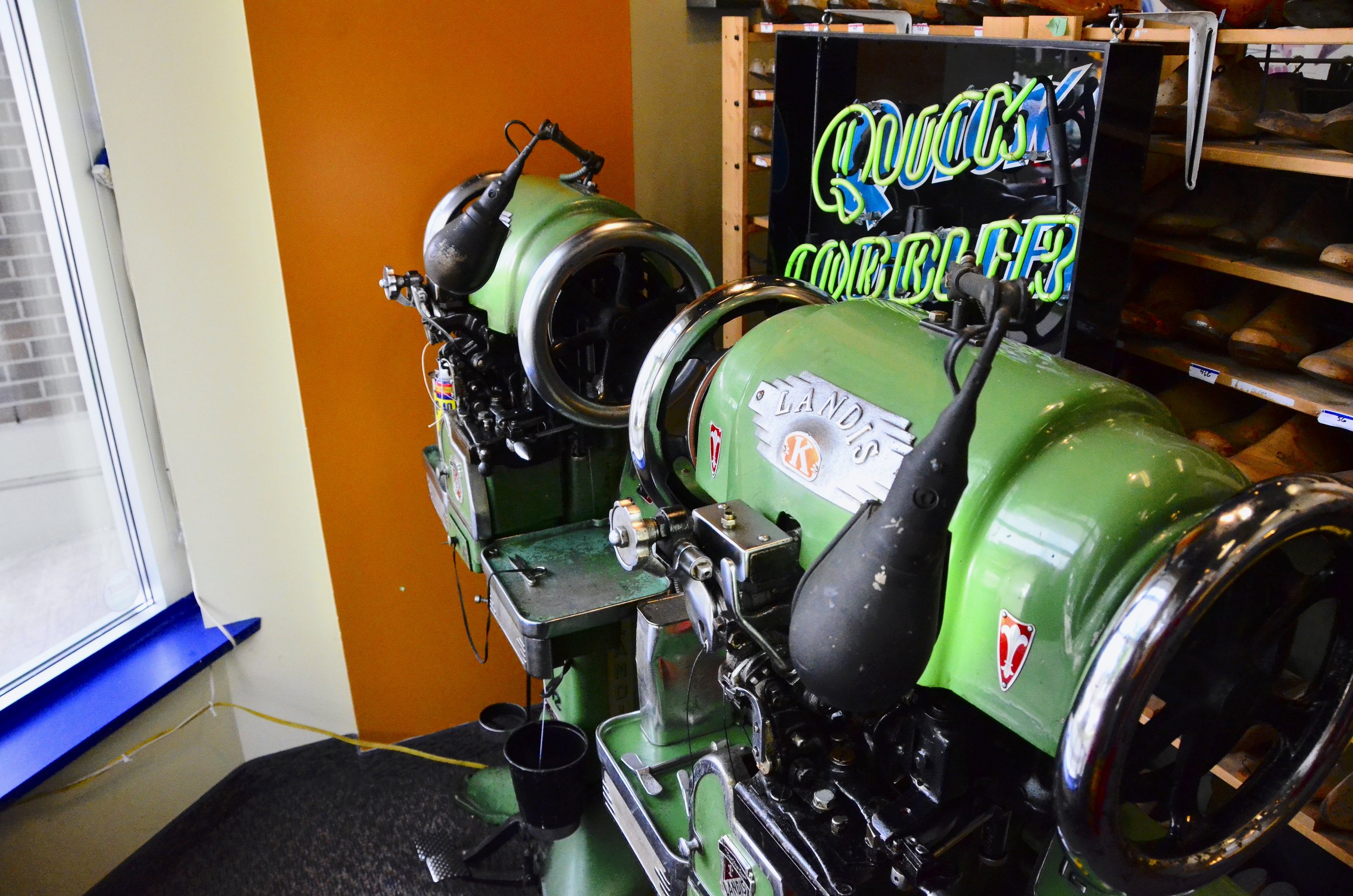 A vintage green machine with the name 'Landis' on a silver nameplate, displayed in a workshop with a neon sign behind it reading 'Touch Cleaner'. The machine has black and silver parts, and is situated near a window and shelves with shoes.